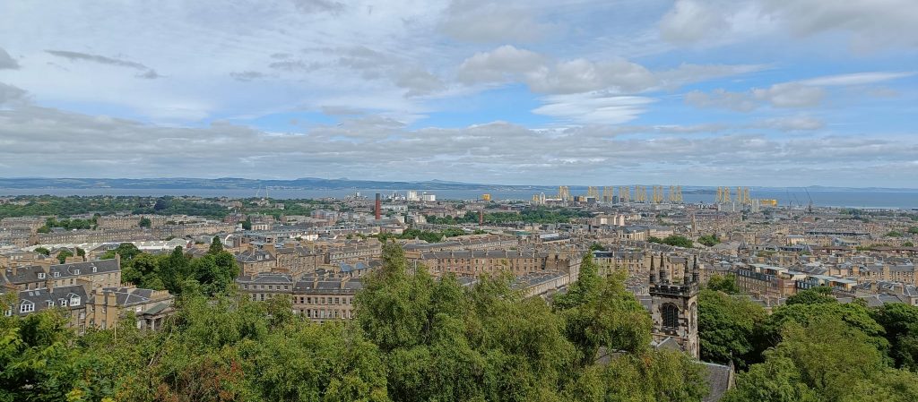 Paronamic photography of the city of Edinburgh on a partial-cloudy day, with tree tops at the bottom, the city at the center of the image and with views of the harbour and the North Sea.